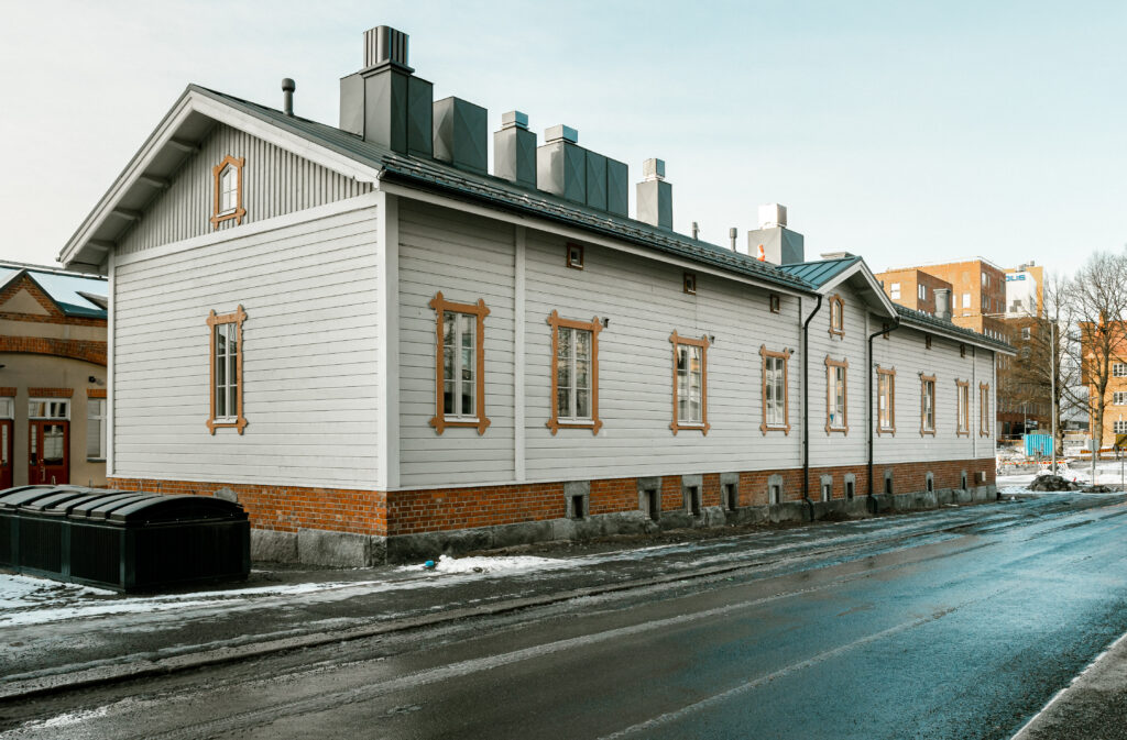 Kuvassa näkyy Tavara-aseman naapurissa sijaitseva vanha valkoinen Puutalo kuvattuna autotieltä päin. Taustalla pilkottavat maitokahvin värinen Tavara-asema ja Tammelan kerrostalot..