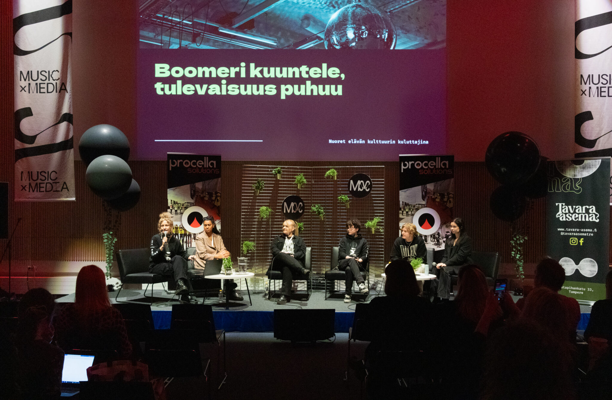 "Boomeri kuuntele, tulevaisuus puhuu" -keskustelun panelistit ja moderaattori istuvat Tampere-talon kokoussalin lavalla. Huone on täynnä väkeä, ja valaistus tilassa on hämärä. Lavan takana näkyy diaesitys, mainosjulisteita ja vihrekasveja.