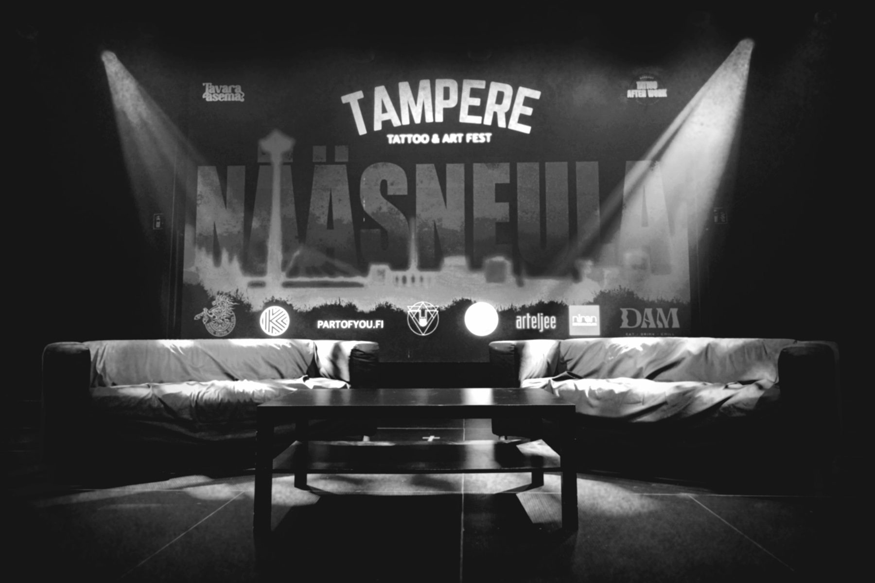 Mustavalkoinen valokuva Tavara-aseman lavalta, jonne on aseteltu kaksi isoa sohvaa ja matala pöytä tapahtuman keskustelijoita varten. Taustalle on heijastettu Tampere Tattoo & Art Festin iso logo ja kuvituskuva. Kaksi valonheitintä valaisee lavaa.